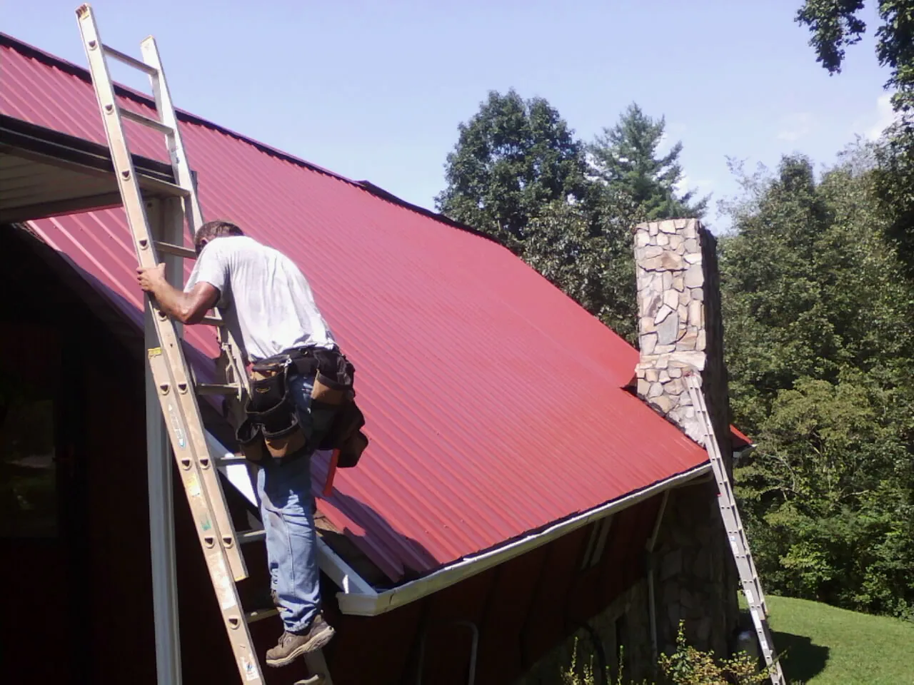 Roofer on a ladder beside a red metal roof and stone chimney — Soffit & Fascia Repair by Roofing Brothers in Milton